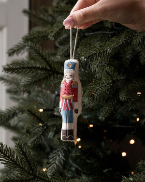 A hand holds a small embroidered nutcracker Christmas ornament against the green branches of a lit Christmas tree.