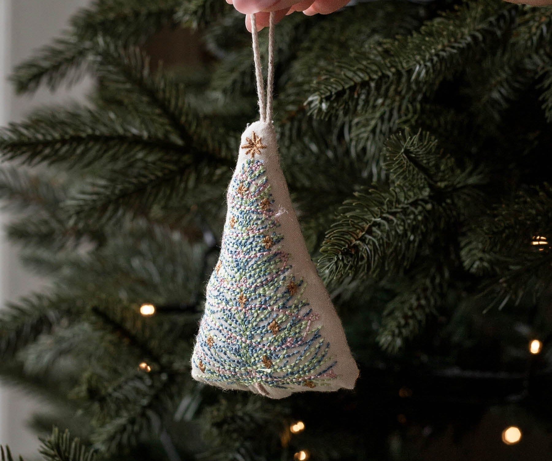 A hand holds a small triangular embroidered Christmas tree ornament with a white string hanger, featuring blue, green, and gold thread details, set against a blurred Christmas tree background with warm lights.
