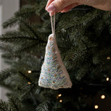 A hand holds a small triangular embroidered Christmas tree ornament with a white string hanger, featuring blue, green, and gold thread details, set against a blurred Christmas tree background with warm lights.