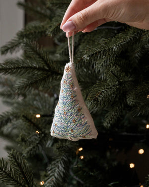 A hand holds a small triangular embroidered Christmas tree ornament with a white string hanger, featuring blue, green, and gold thread details, set against a blurred Christmas tree background with warm lights.