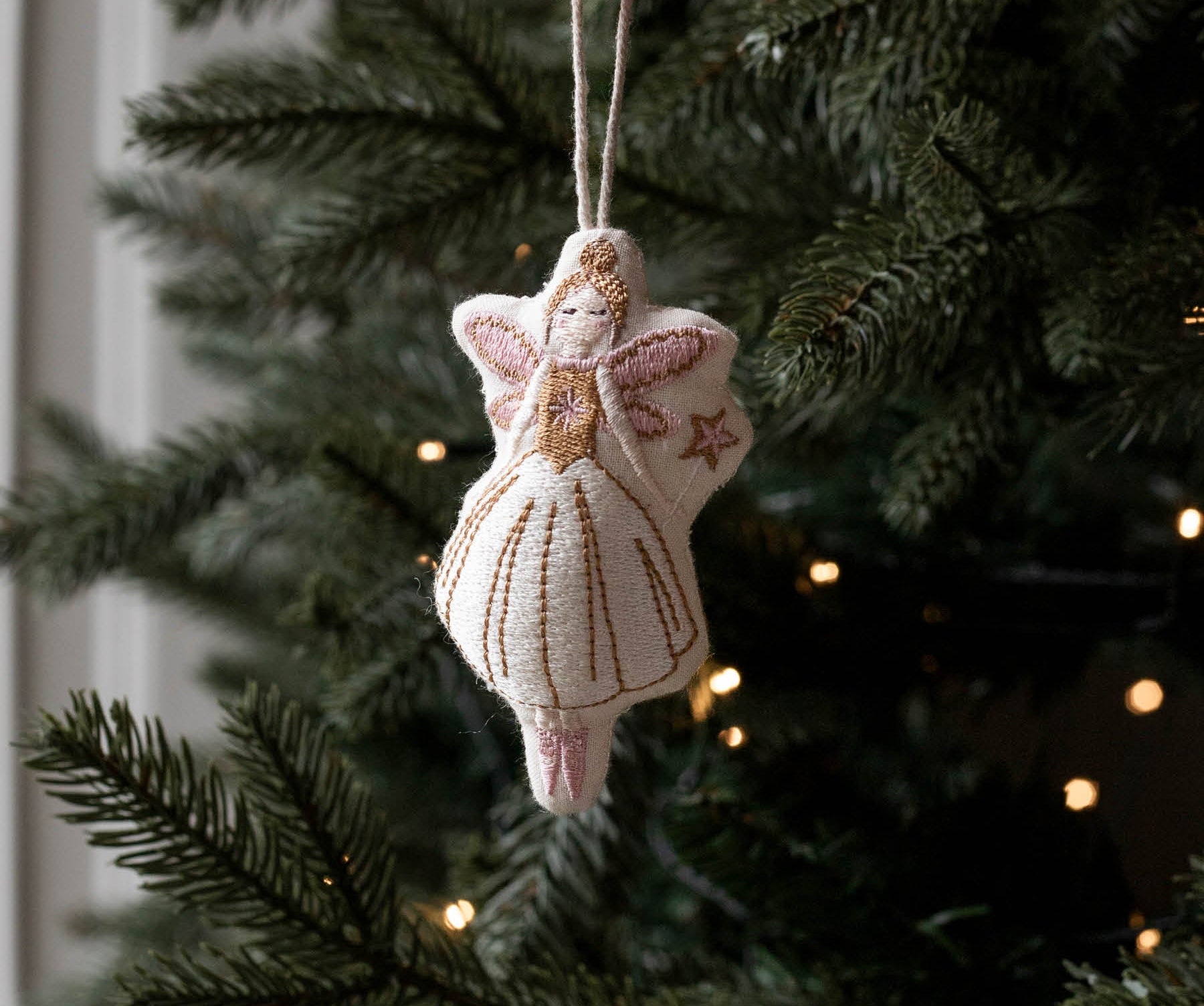 A hand holds a small handmade felt Christmas angel ornament with a white dress and gold embroidery, hanging against a dark green Christmas tree with warm string lights.