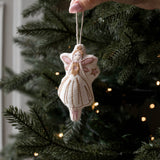 A hand holds a small handmade felt Christmas angel ornament with a white dress and gold embroidery, hanging against a dark green Christmas tree with warm string lights.