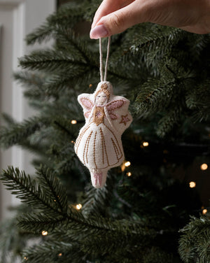 A hand holds a small handmade felt Christmas angel ornament with a white dress and gold embroidery, hanging against a dark green Christmas tree with warm string lights.