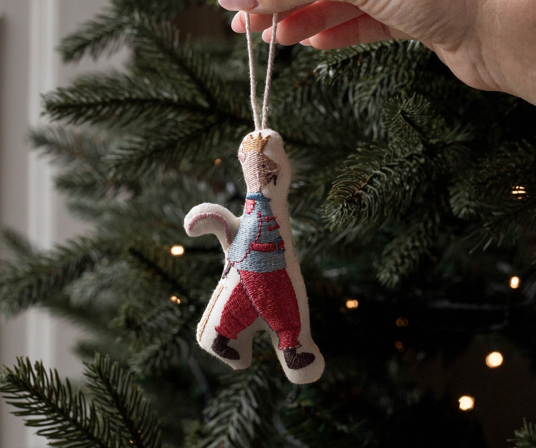 A hand hanging a small handmade felt Christmas ornament shaped like a person in a striped sweater and red pants on a decorated Christmas tree.