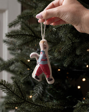 A hand hanging a small handmade felt Christmas ornament shaped like a person in a striped sweater and red pants on a decorated Christmas tree.