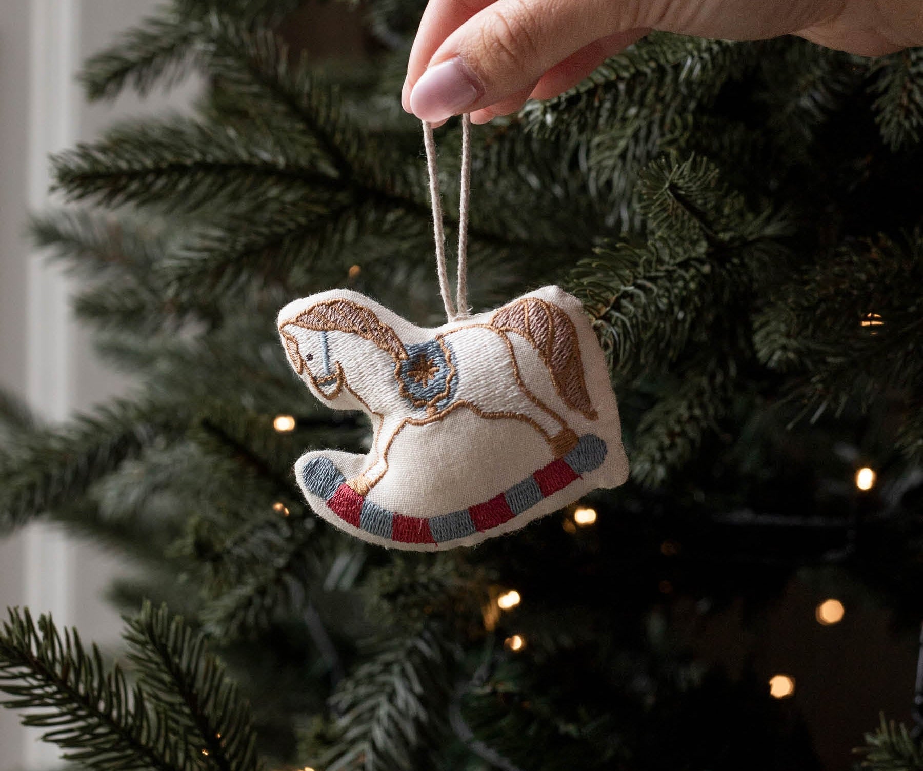 A hand with pink nail polish hangs a small embroidered rocking horse ornament from a string in front of a decorated Christmas tree with warm lights.