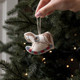 A hand with pink nail polish hangs a small embroidered rocking horse ornament from a string in front of a decorated Christmas tree with warm lights.