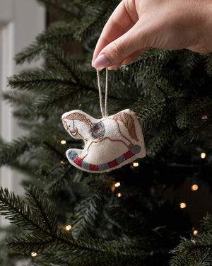 A hand with pink nail polish hangs a small embroidered rocking horse ornament from a string in front of a decorated Christmas tree with warm lights.