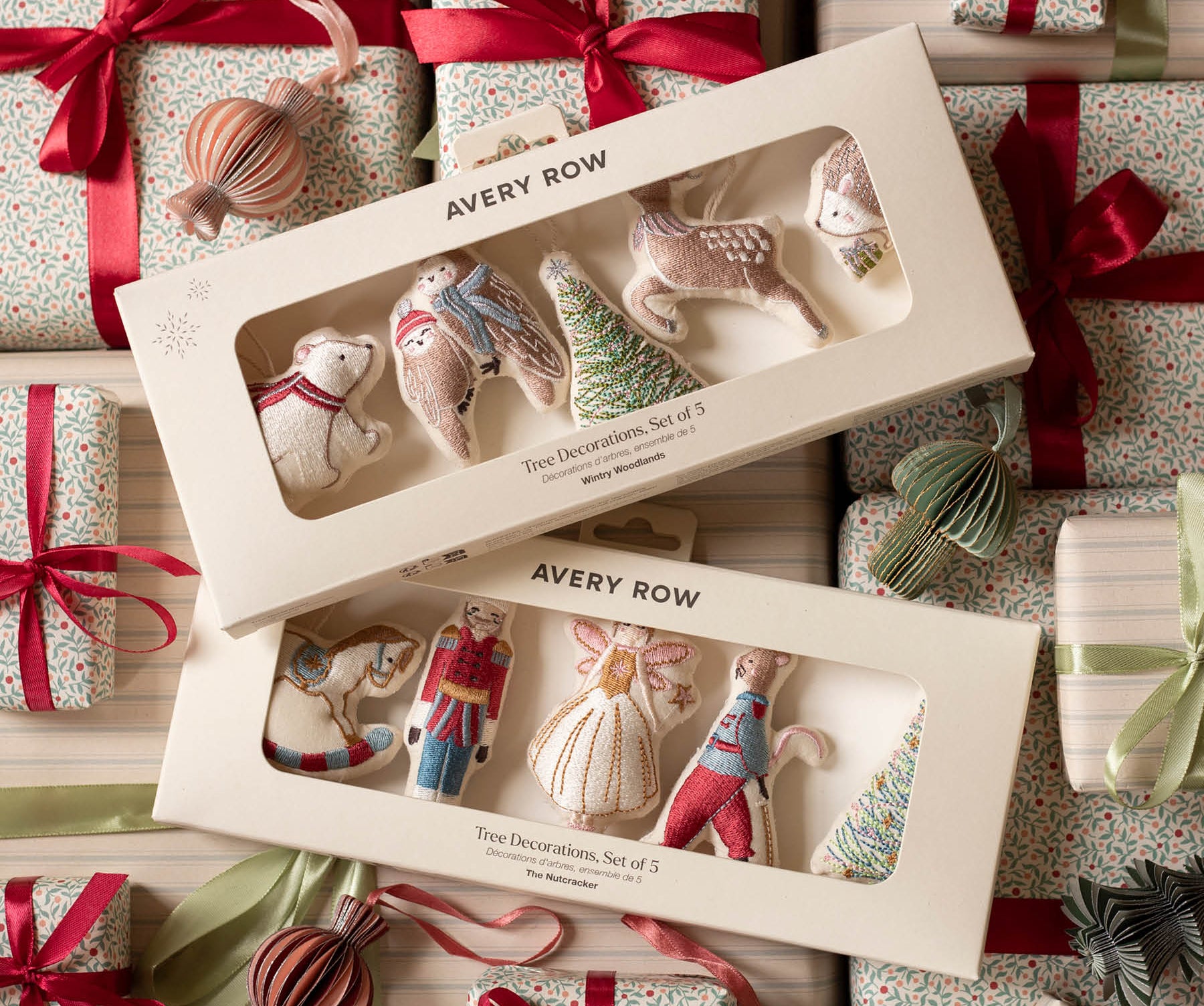 Two boxed sets of Avery Row Christmas tree decorations arranged on top of wrapped gift boxes with red ribbons and ornaments. The boxes contain small fabric holiday ornaments shaped like a bear, bird, tree, and other festive forms, visible through clear window packaging.