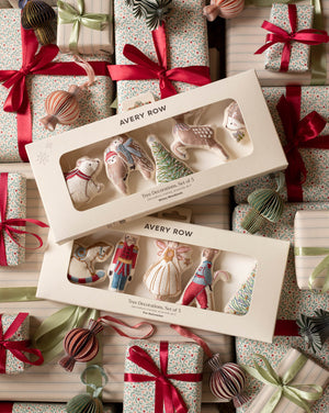Two boxed sets of Avery Row Christmas tree decorations arranged on top of wrapped gift boxes with red ribbons and ornaments. The boxes contain small fabric holiday ornaments shaped like a bear, bird, tree, and other festive forms, visible through clear window packaging.
