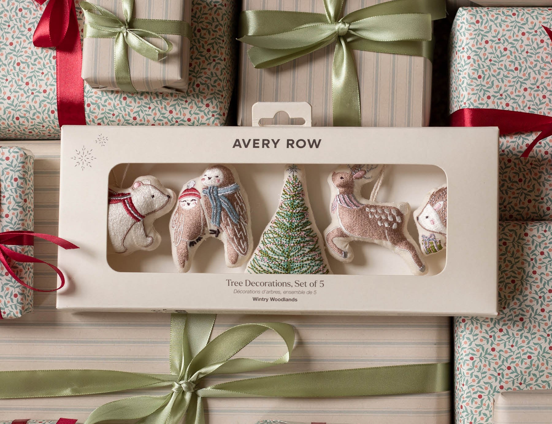 Avery Row Christmas tree decorations set of 5 in a cream-colored gift box, displayed on top of wrapped gift boxes with red ribbons and green satin bows.