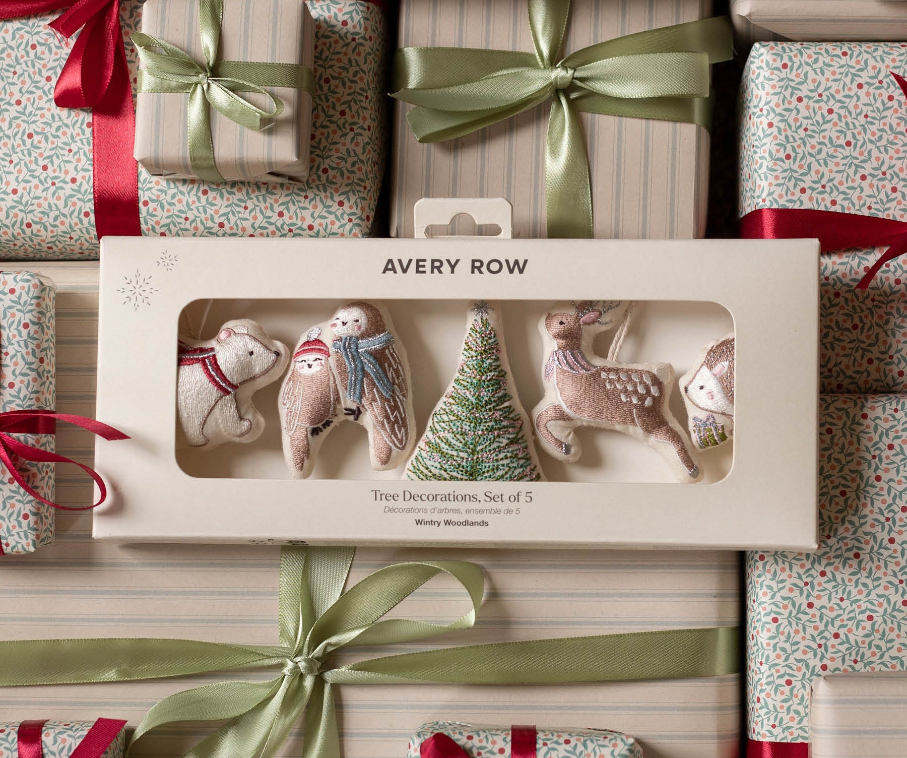 Avery Row Christmas tree decorations set of 5 in a cream-colored gift box, displayed on top of wrapped gift boxes with red ribbons and green satin bows.