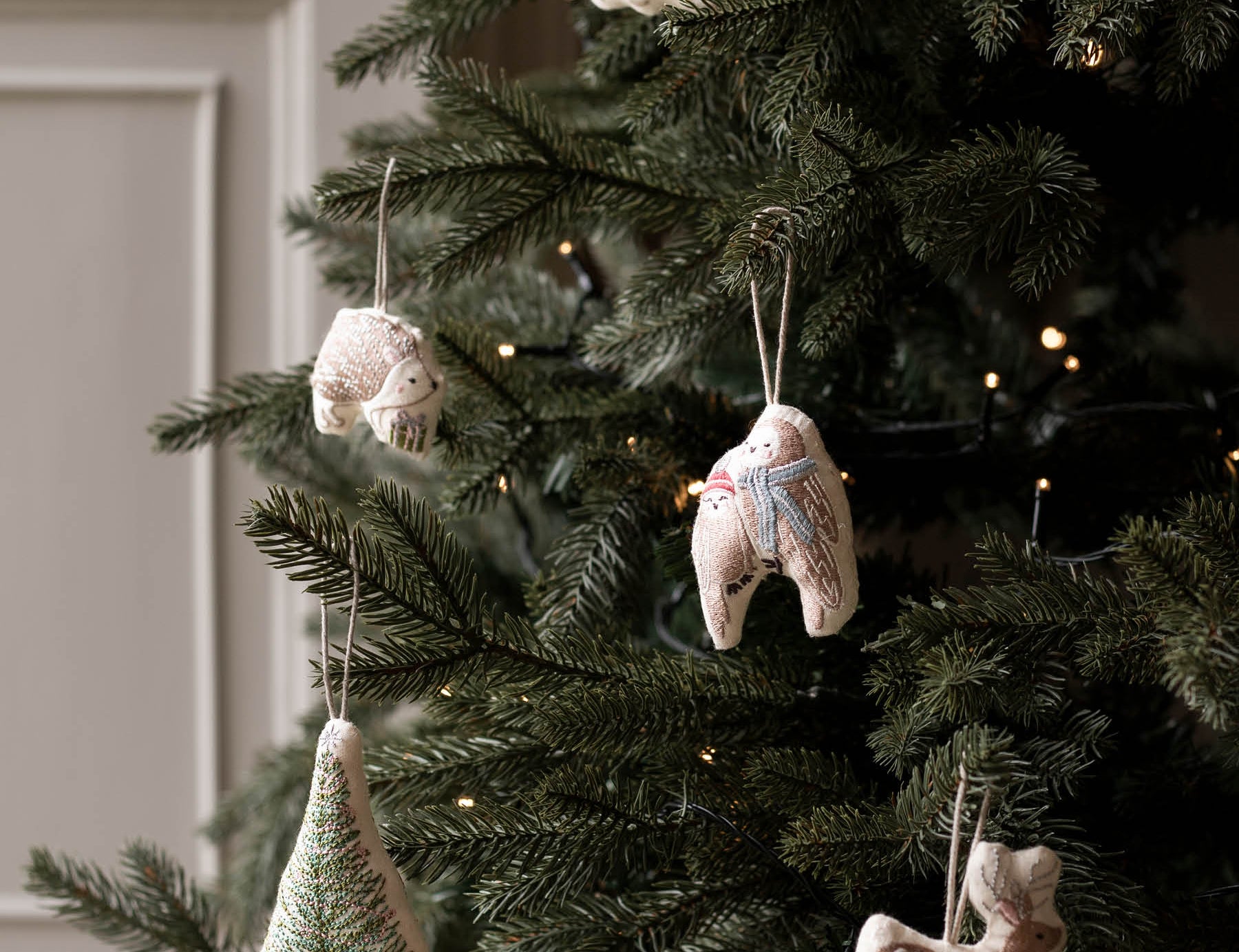 Handmade ceramic Christmas ornaments hanging on a decorated Christmas tree, including a tree-shaped ornament, a snowman, a rabbit, and a deer, with warm string lights in the background.