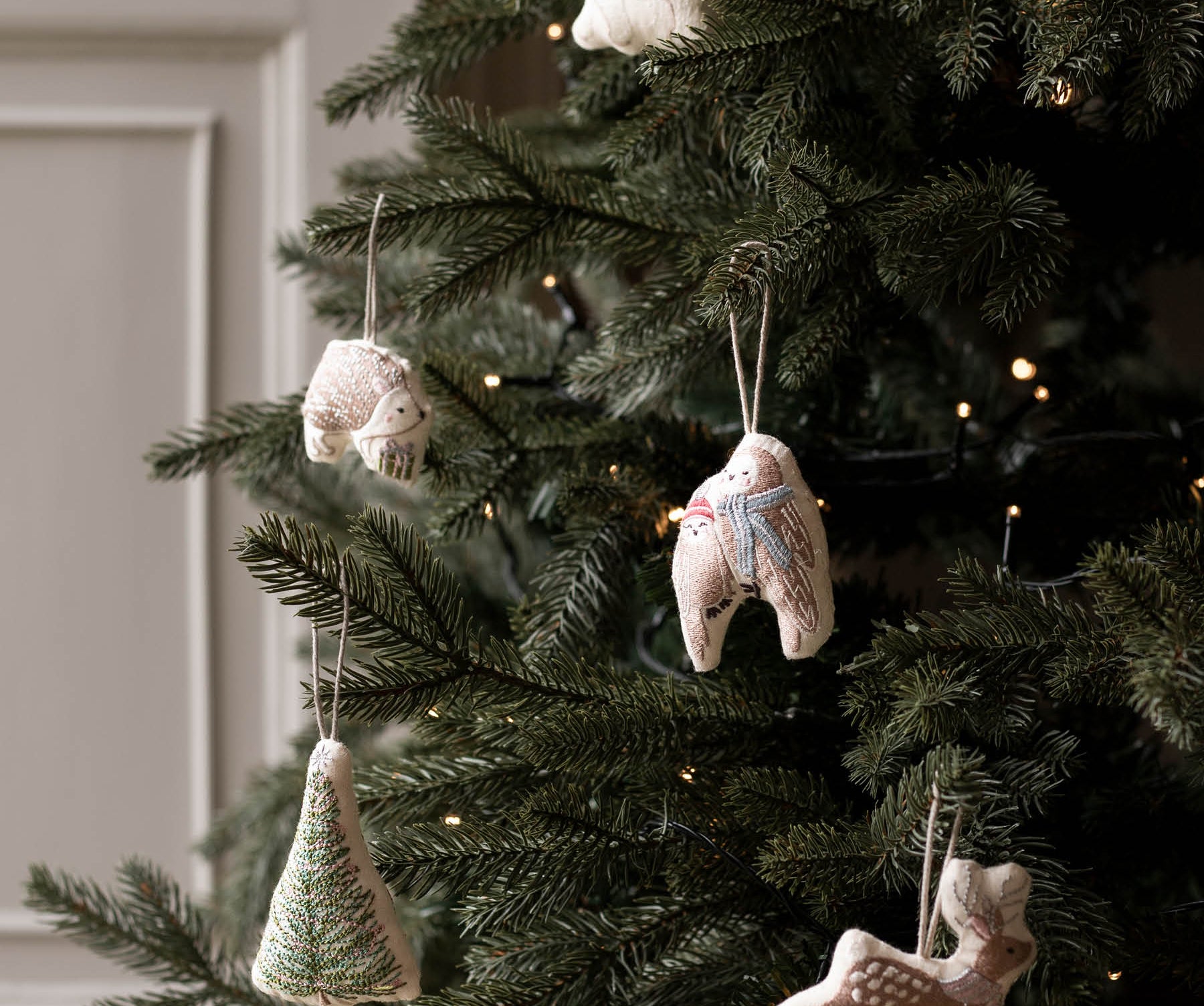 Handmade ceramic Christmas ornaments hanging on a decorated Christmas tree, including a tree-shaped ornament, a snowman, a rabbit, and a deer, with warm string lights in the background.