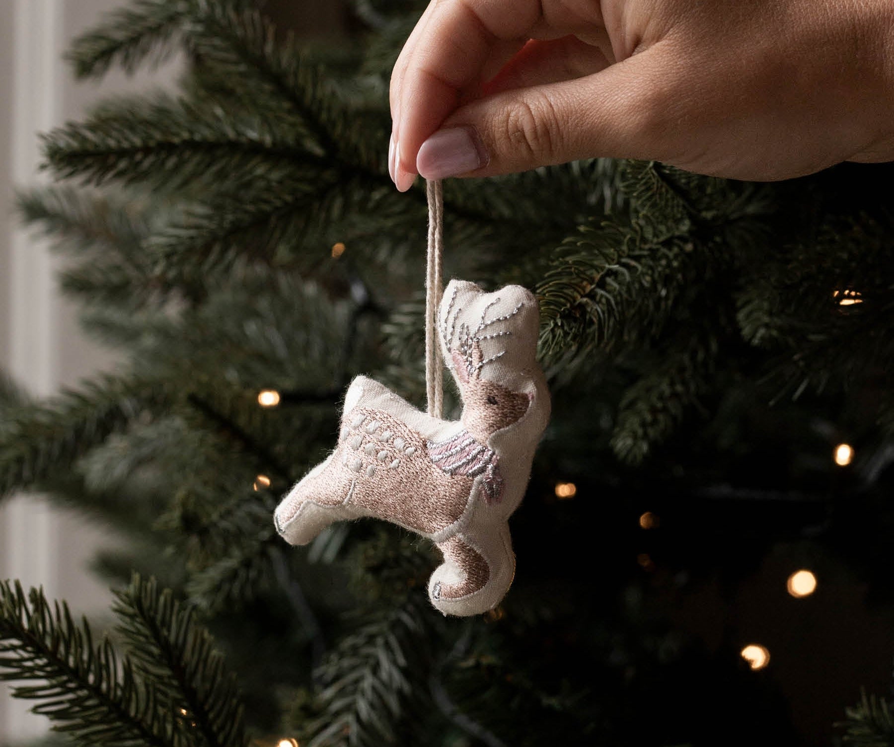 A hand hangs a small beige embroidered angel ornament from a green Christmas tree with warm string lights in the background.