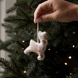 A hand hangs a small beige embroidered angel ornament from a green Christmas tree with warm string lights in the background.