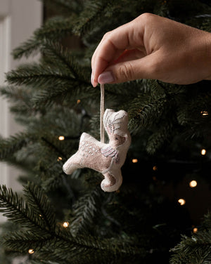A hand hangs a small beige embroidered angel ornament from a green Christmas tree with warm string lights in the background.