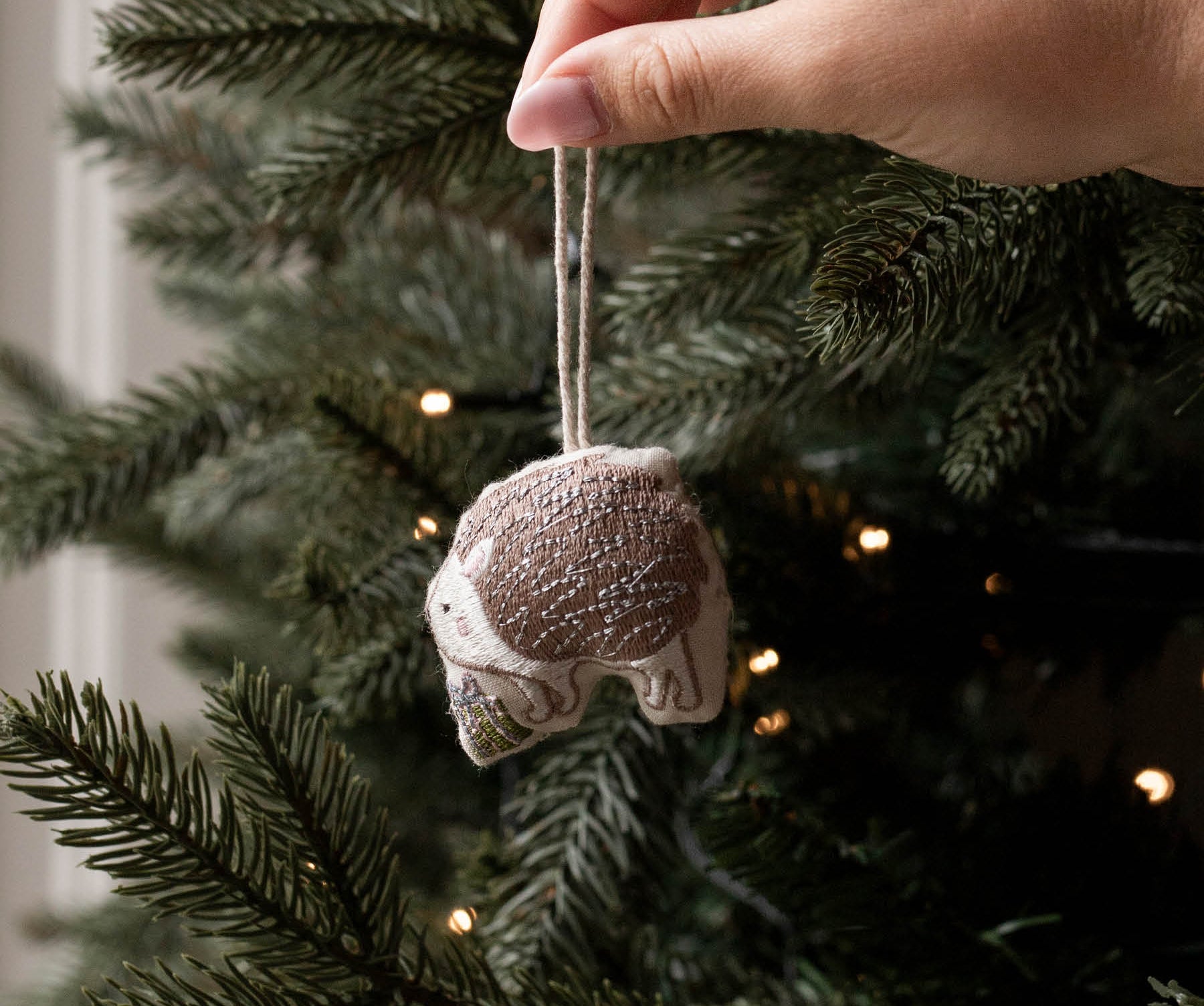 A hand hanging a small fabric ornament shaped like a sleeping cat from a decorated Christmas tree. The ornament is beige and brown with a white string, shown against the green pine branches and warm string lights of the tree.
