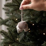 A hand hanging a small fabric ornament shaped like a sleeping cat from a decorated Christmas tree. The ornament is beige and brown with a white string, shown against the green pine branches and warm string lights of the tree.