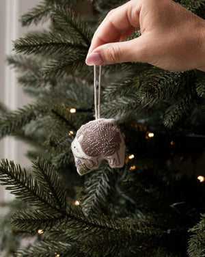 A hand hanging a small fabric ornament shaped like a sleeping cat from a decorated Christmas tree. The ornament is beige and brown with a white string, shown against the green pine branches and warm string lights of the tree.