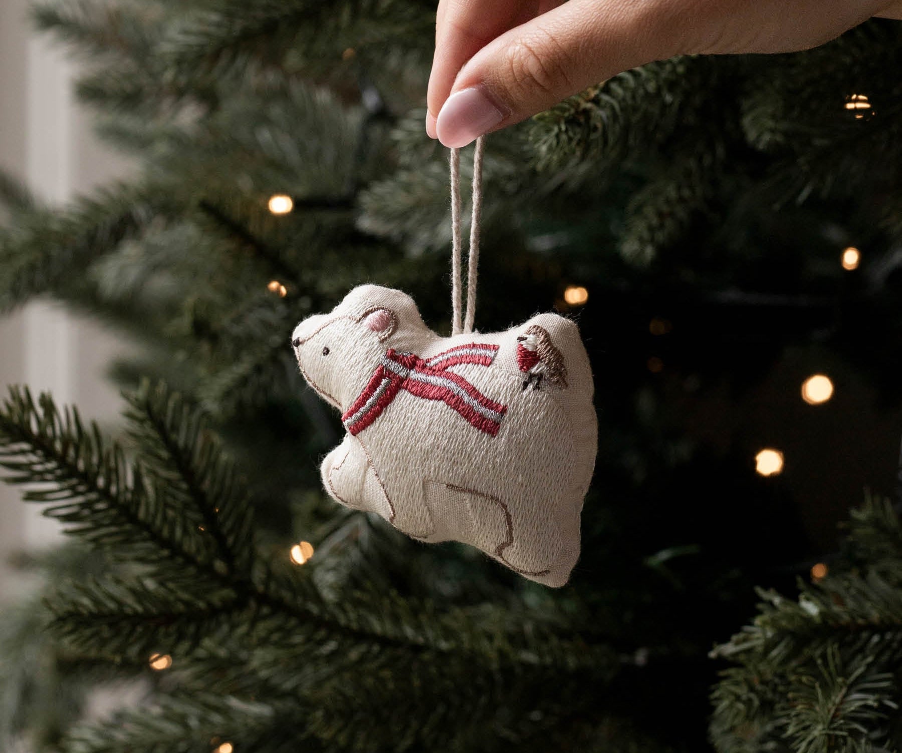 A hand hangs a small beige fabric ornament shaped like a polar bear wearing a red striped scarf from a dark green Christmas tree with warm string lights.
