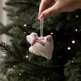 A hand hangs a small beige fabric ornament shaped like a polar bear wearing a red striped scarf from a dark green Christmas tree with warm string lights.