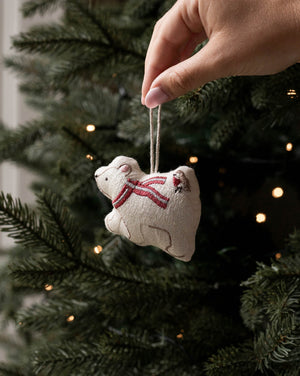 A hand hangs a small beige fabric ornament shaped like a polar bear wearing a red striped scarf from a dark green Christmas tree with warm string lights.