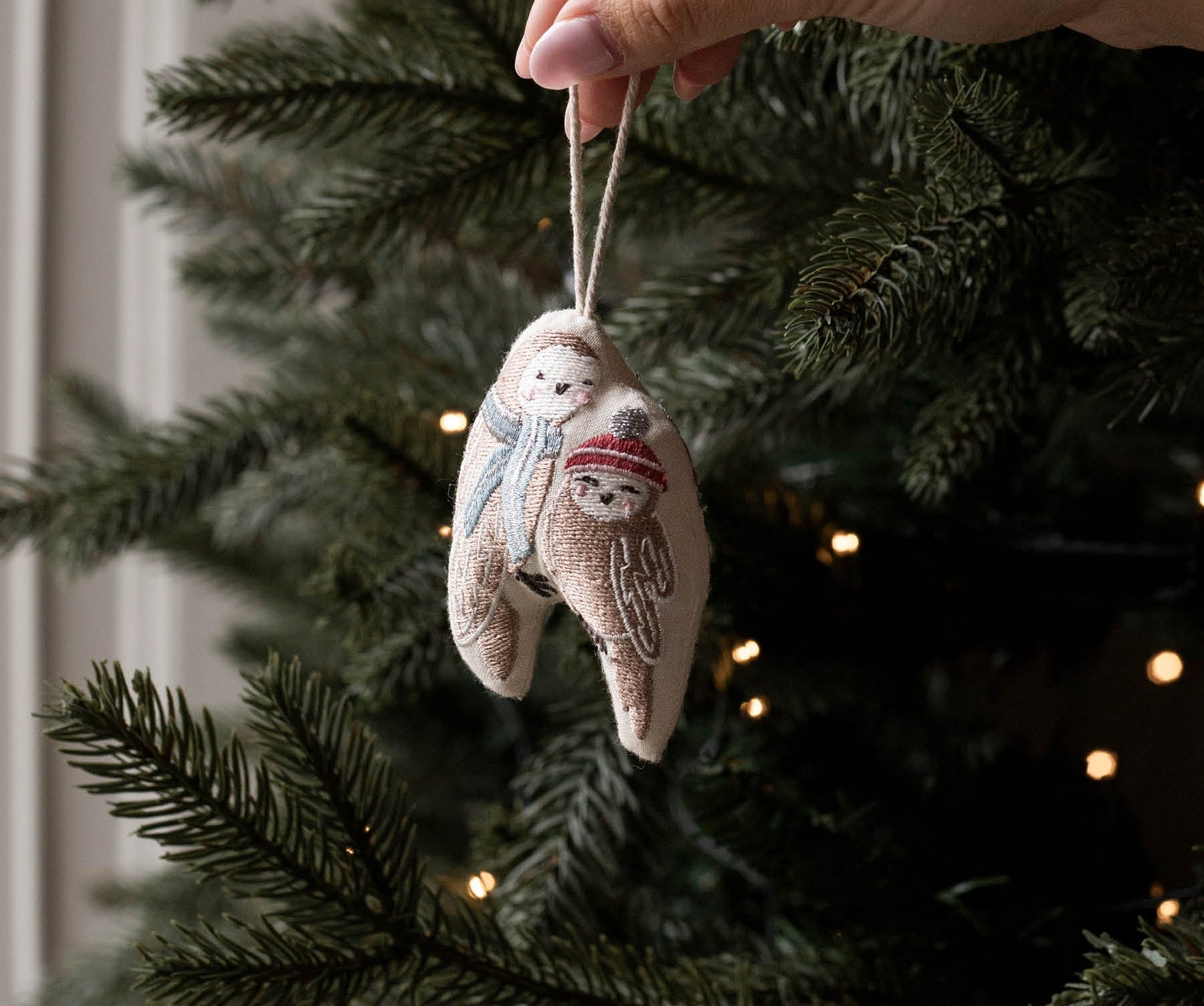 A hand hangs a small embroidered fabric ornament shaped like a snowman on a Christmas tree. The ornament is beige with white embroidery and a red scarf, with the tree branches and warm string lights blurred in the background.
