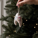A hand hangs a small embroidered fabric ornament shaped like a snowman on a Christmas tree. The ornament is beige with white embroidery and a red scarf, with the tree branches and warm string lights blurred in the background.