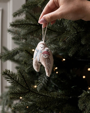 A hand hangs a small embroidered fabric ornament shaped like a snowman on a Christmas tree. The ornament is beige with white embroidery and a red scarf, with the tree branches and warm string lights blurred in the background.
