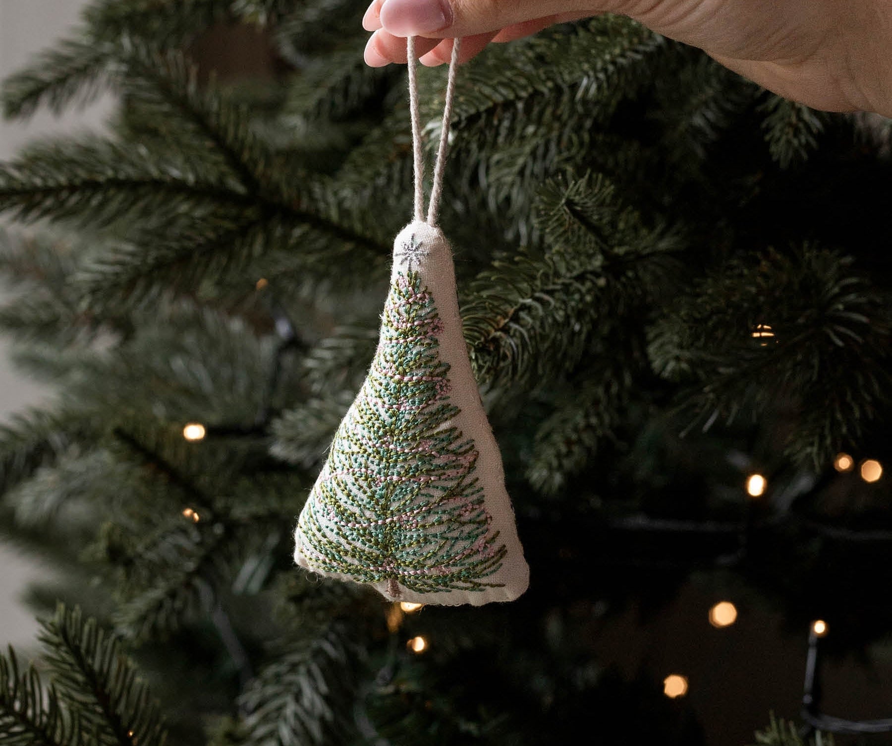 A hand holds a small triangular fabric Christmas tree ornament with green and white embroidered branches, hanging in front of a real Christmas tree with warm string lights.