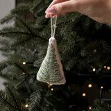 A hand holds a small triangular fabric Christmas tree ornament with green and white embroidered branches, hanging in front of a real Christmas tree with warm string lights.