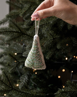 A hand holds a small triangular fabric Christmas tree ornament with green and white embroidered branches, hanging in front of a real Christmas tree with warm string lights.