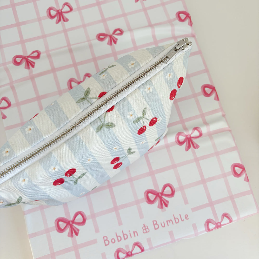 Nappy Wallet Cherry Stripe, seen from above with zip facing up, lying on a pink bow gingham mat