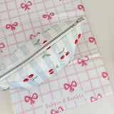 Nappy Wallet Cherry Stripe, seen from above with zip facing up, lying on a pink bow gingham mat