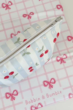 Nappy Wallet Cherry Stripe, seen from above with zip facing up, lying on a pink bow gingham mat