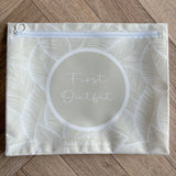 Hospital bag organiser pouches, beige leaf, flat pouch labelled First Outfit on a wooden floor