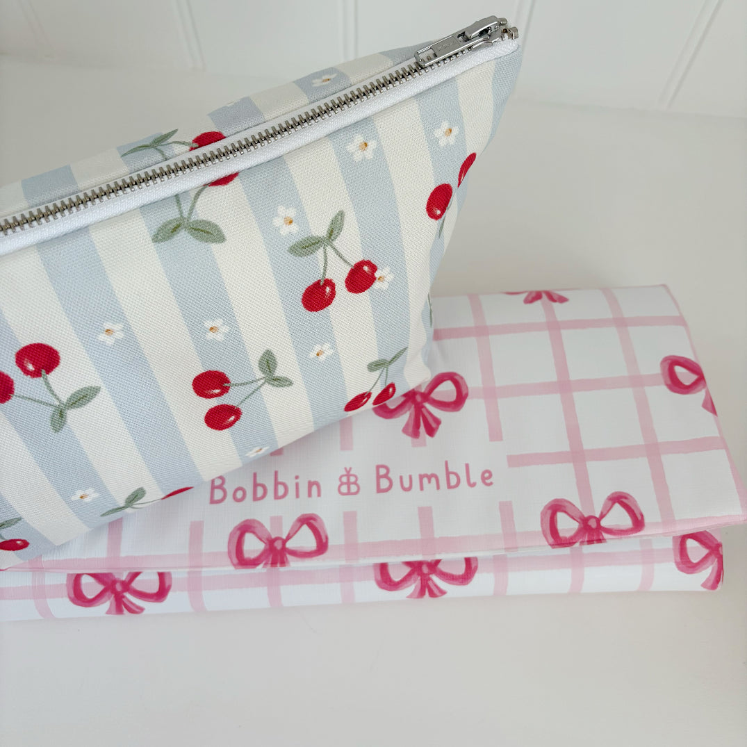 Nappy Wallet Cherry Stripe, resting on a folded pink gingham changing mat with bows labelled Bobbin and Bumble