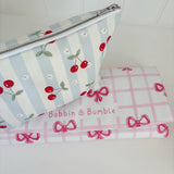 Nappy Wallet Cherry Stripe, resting on a folded pink gingham changing mat with bows labelled Bobbin and Bumble
