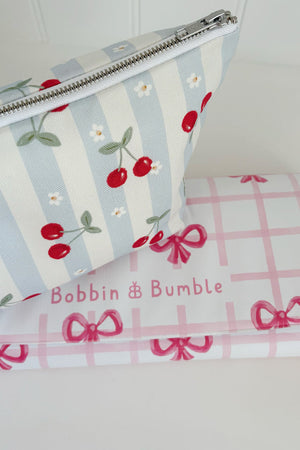Nappy Wallet Cherry Stripe, resting on a folded pink gingham changing mat with bows labelled Bobbin and Bumble
