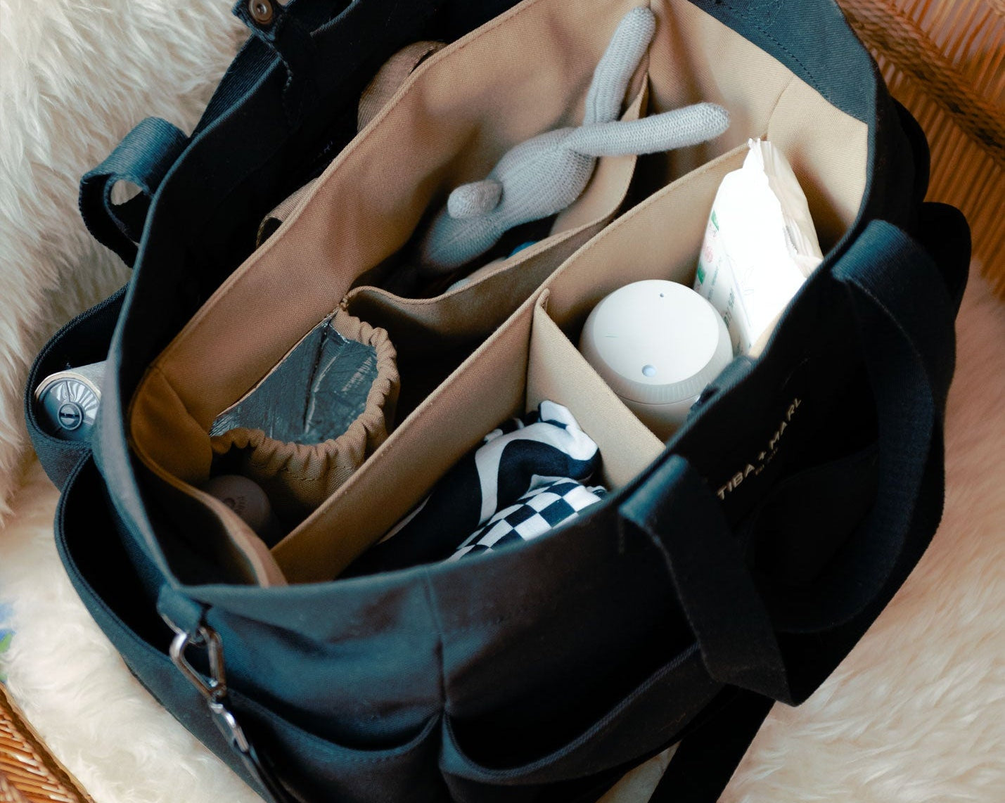 Black diaper bag with compartments containing baby items on a fluffy surface
