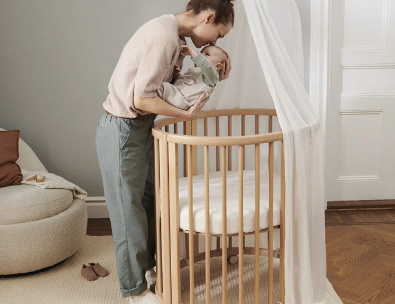 Stokke Sleepi Canopy white above a round cot as a woman leans in to kiss the baby she is holding