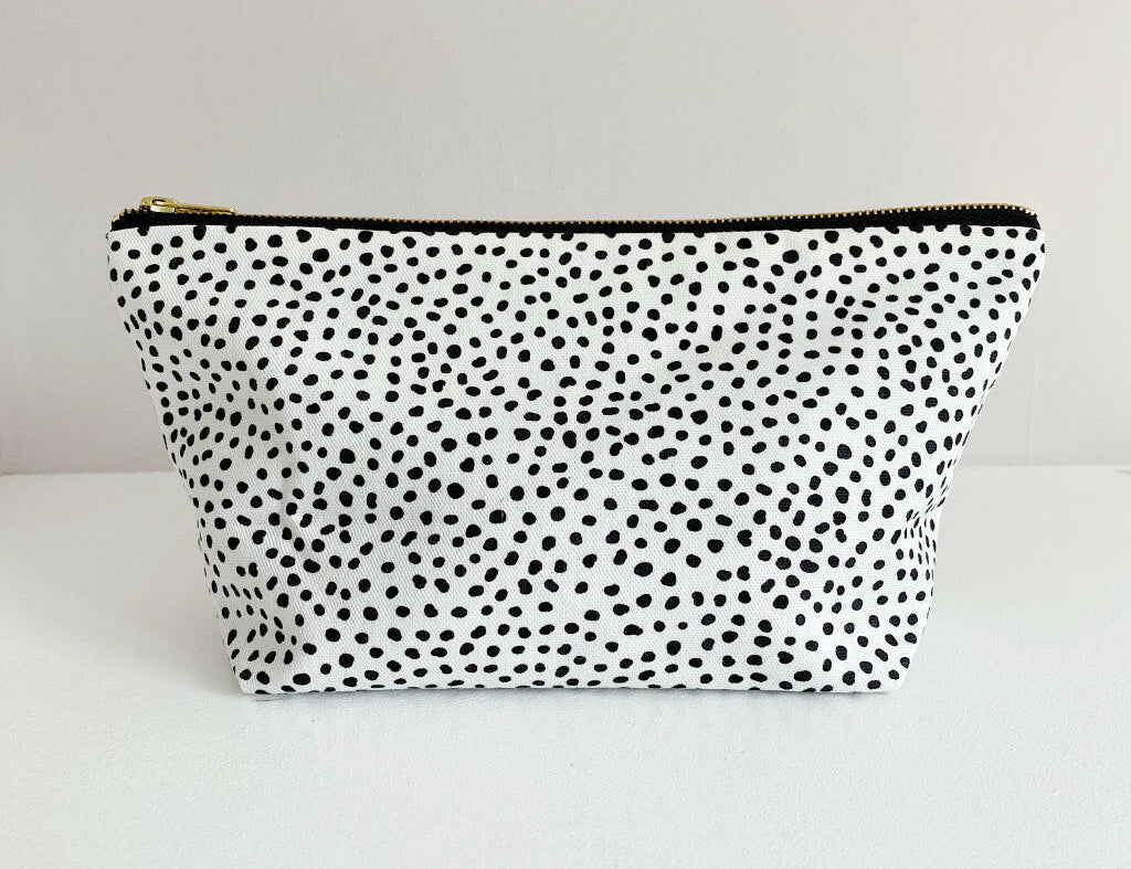 Nappy wallet in dalmatian print, front view of the black spotted white pouch with a gold zip on a plain surface