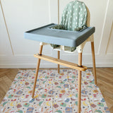 Splash Mat Snack Time laid on a wooden floor under a blue-grey highchair with a bamboo footrest