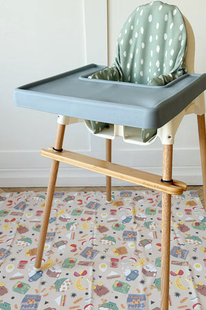 Splash Mat Snack Time laid on a wooden floor under a blue-grey highchair with a bamboo footrest