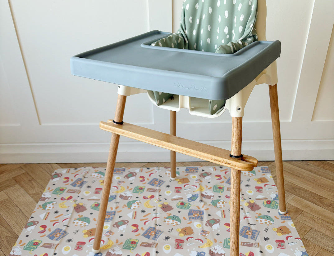 Splash Mat Snack Time laid on a wooden floor under a blue-grey highchair with a bamboo footrest