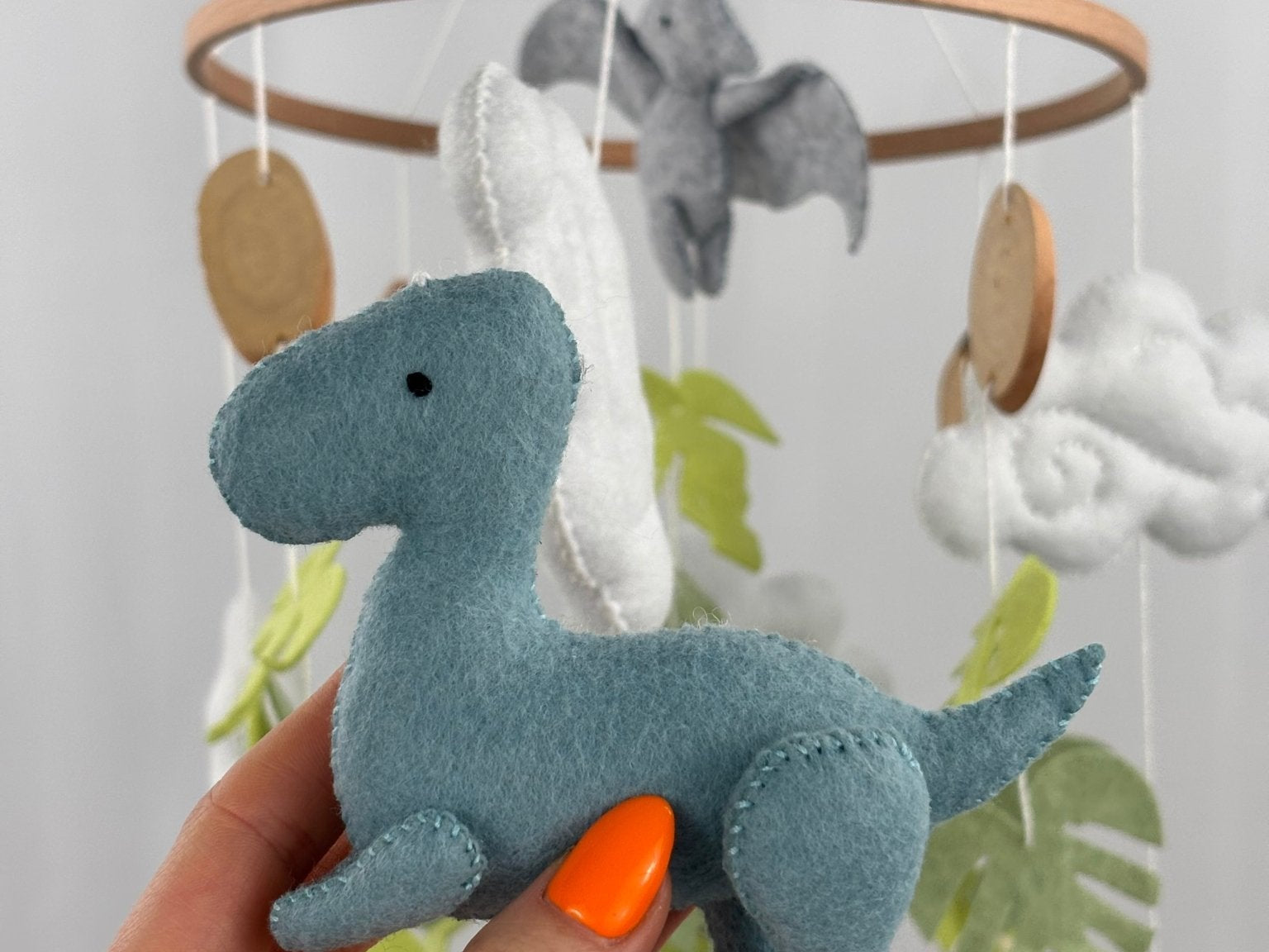 Little Dino Land felt cot mobile, hand holding the teal dinosaur in side view with a small black eye