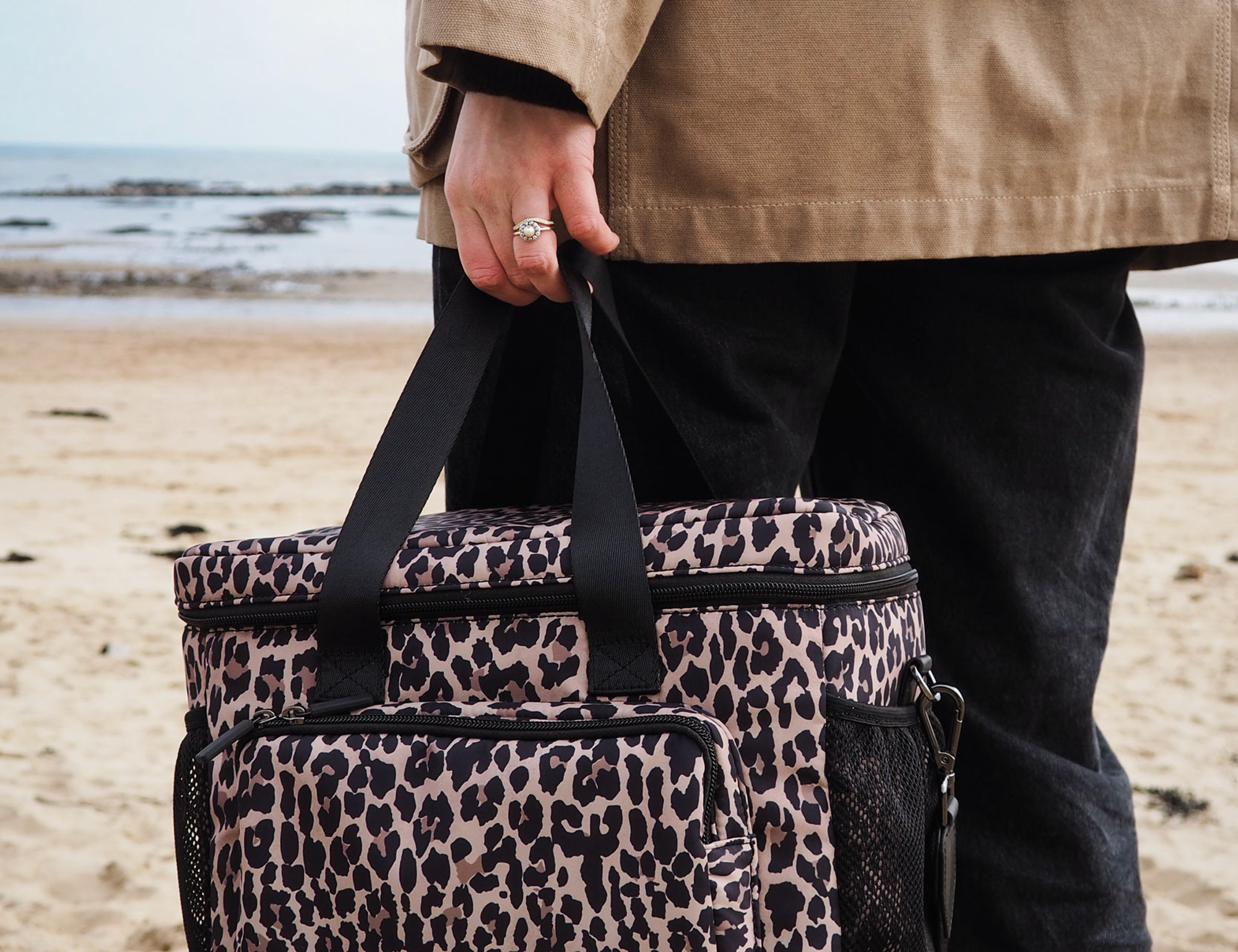 T+M Family Picnic Cool Bag in Lighter Leopard Print, held by its handles by a person on a beach