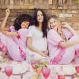 Three women sitting together on a pink and white striped bench in front of a brick wall, wearing matching pink and white ruffled sleepwear sets. One woman wears a white top with white bottoms, while the other two wear light pink and medium pink ruffled robes with matching pink ruffled shorts.