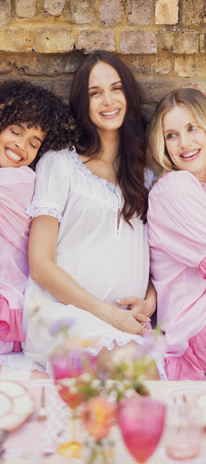 Three women sitting together on a pink and white striped bench in front of a brick wall, wearing matching pink and white ruffled sleepwear sets. One woman wears a white top with white bottoms, while the other two wear light pink and medium pink ruffled robes with matching pink ruffled shorts.